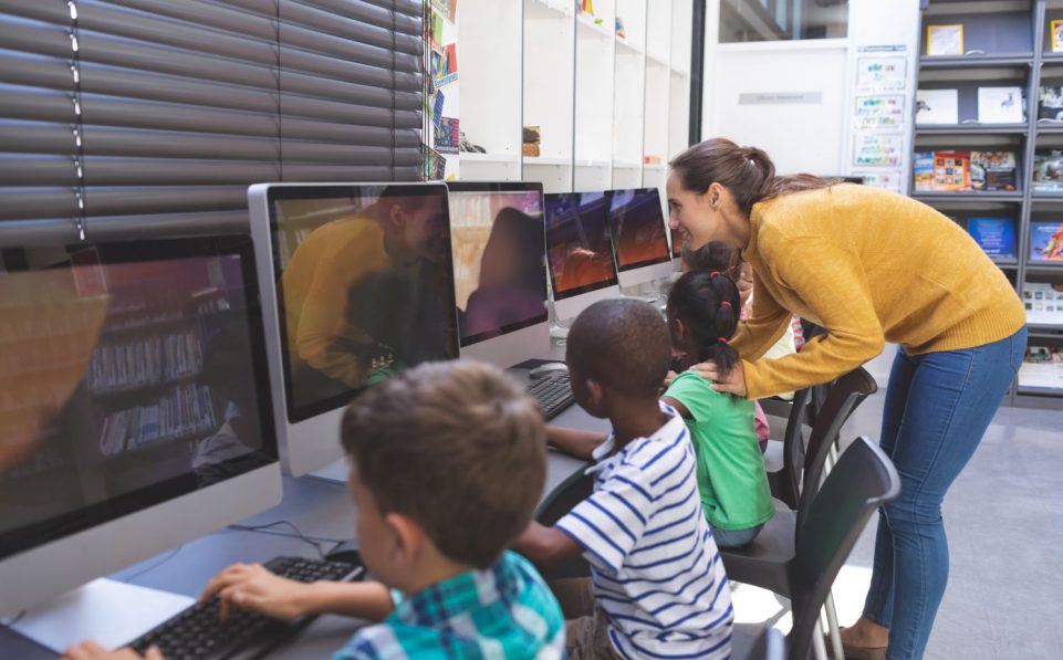 Side view of happy caucasian teacher teaching African-american schoolgirl while others students working on computers  in computer room